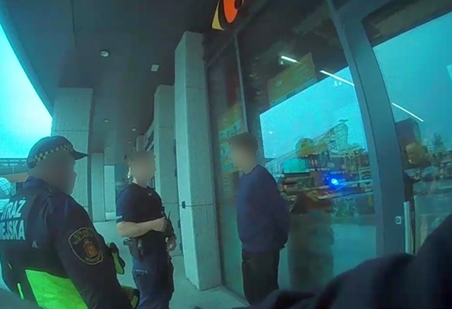 Dwaj policjanci rozmawiają z mężczyzną w niebieskiej bluzie przed sklepem, w tle widać atrakcje w wesołym miasteczku.