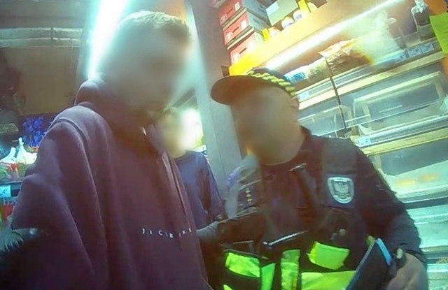 Mężczyzna w bluzie rozmawia z policjantem w sklepie spożywczym, w tle widoczne półki z produktami.