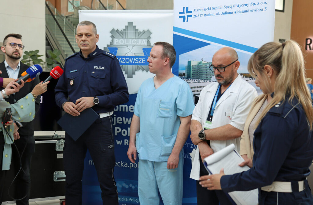 Policjant w mundurze przemawia podczas konferencji prasowej, obok niego lekarze w odzieży medycznej.