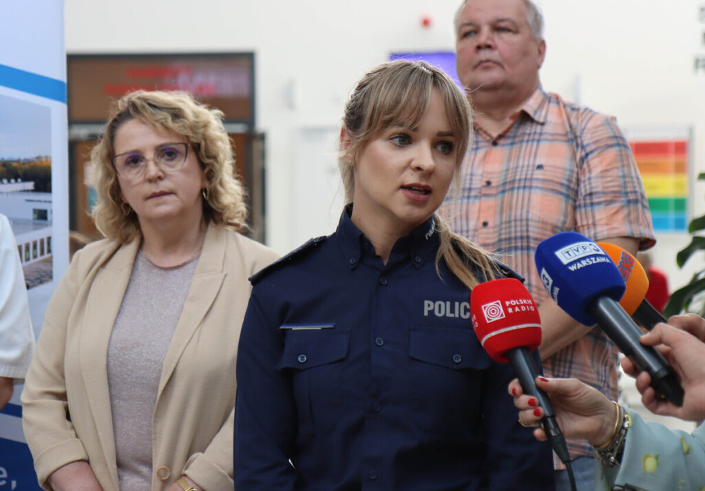 Kobieta w mundurze policyjnym udziela wywiadu, stojąc przed grupą dziennikarzy z mikrofonami. W tle widoczna reklama.