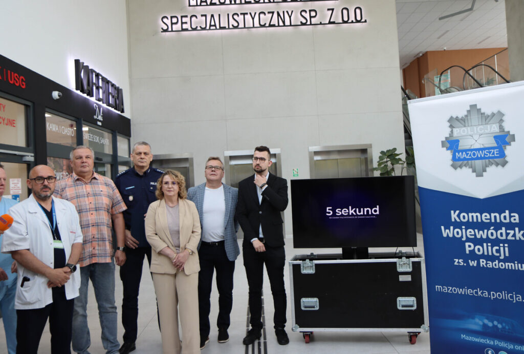 Grupa osób w różnych strojach stoi przed wejściem do szpitala, obok telewizora z napisem "5 sekund".