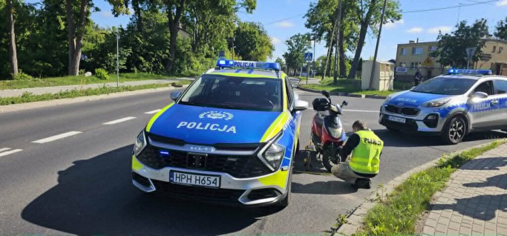Policjant w kamizelce odblaskowej sprawdza motorower przy drodze, obok stoi radiowóz policyjny z włączonymi sygnalizatorami.