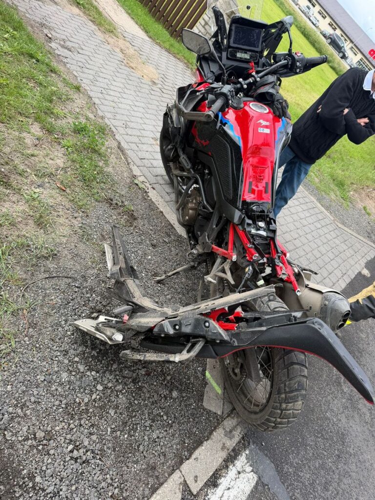 Motocykl w kolorze czerwonym z uszkodzoną tylną częścią stoi na utwardzonej nawierzchni, obok mężczyzna z założonymi rękami.
