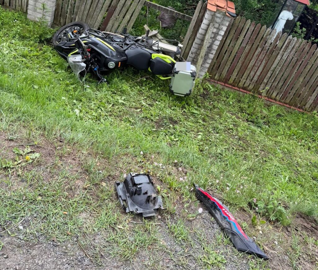 Motocykl leży na boku w trawie obok uszkodzonych elementów, w tle drewniane ogrodzenie.