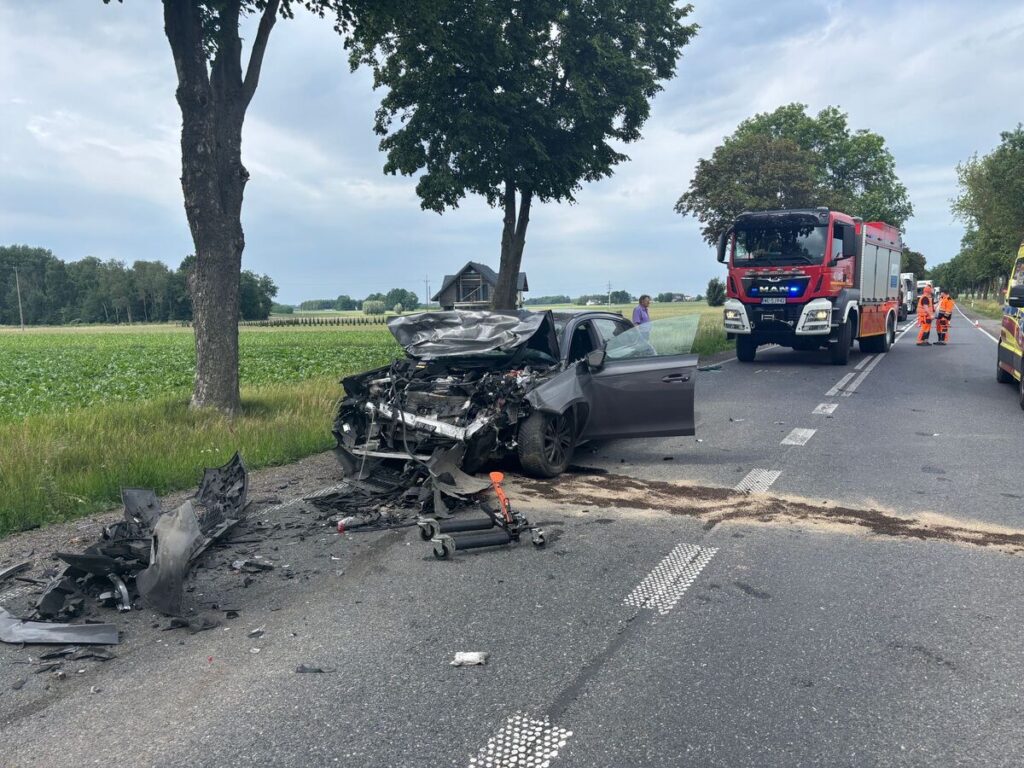 Zniszczony samochód osobowy na poboczu drogi, obok działania służb ratunkowych w pomarańczowych uniformach.