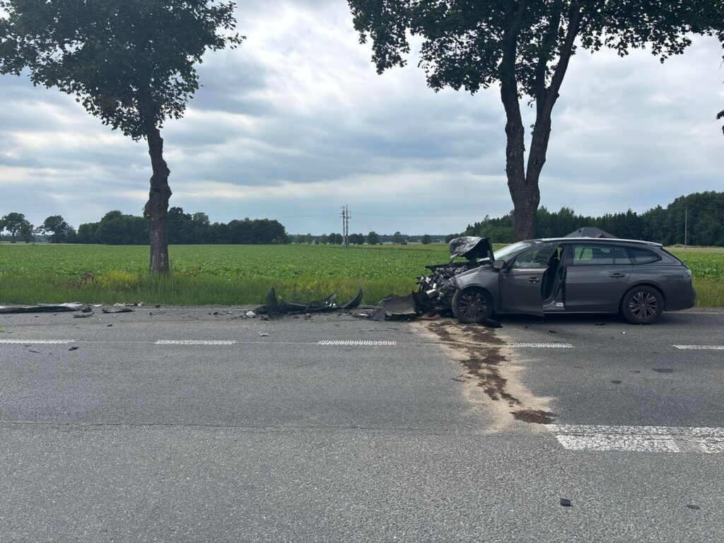 Zniszczony samochód osobowy stoi na drodze, obok leżą fragmenty karoserii, a w tle rozciągają się pola.