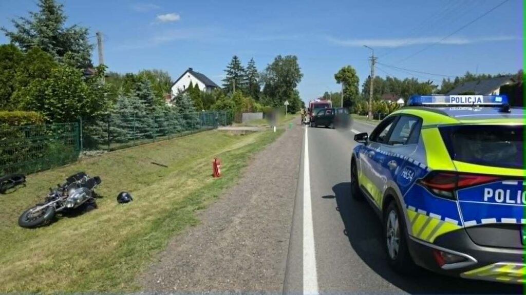 Motocykl leży na trawie obok drogi, a obok niego stoi radiowóz policyjny. W tle widoczne są drzewa i domy.