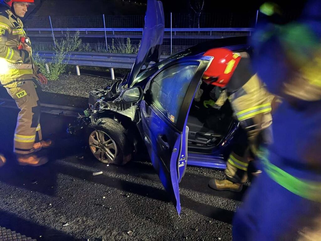 Strażacy w hełmach i mundurach oceniają uszkodzone auto po wypadku na drodze nocą.