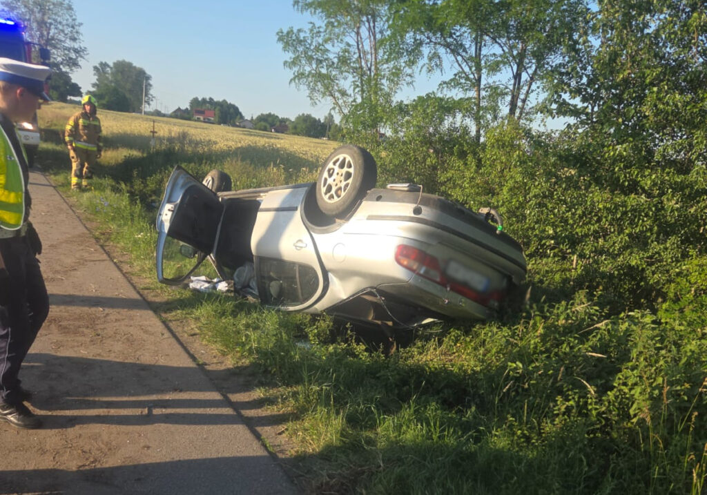 Osobowy samochód leży na dachu na poboczu drogi, obok stoją policjant i strażak, w tle widać pola.