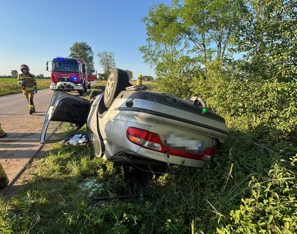 Samochód leżący na dachu w zaroślach przy drodze, obok strażacy interweniujący w akcji ratunkowej.