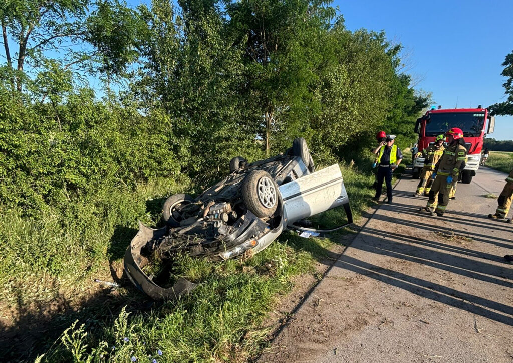 Przewrócony samochód osobowy leży na boku przy drodze, obok grupy strażaków i ratowników medycznych.