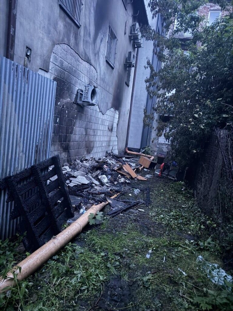 Zniszczona ściana budynku z widocznymi śladami po pożarze, obok gruz i zniszczone meble na zaniedbanym terenie.