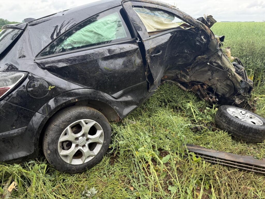 Czarny samochód osobowy leży na boku w zaroślach, z uszkodzeniem nadwozia i jednym odłączonym kołem.