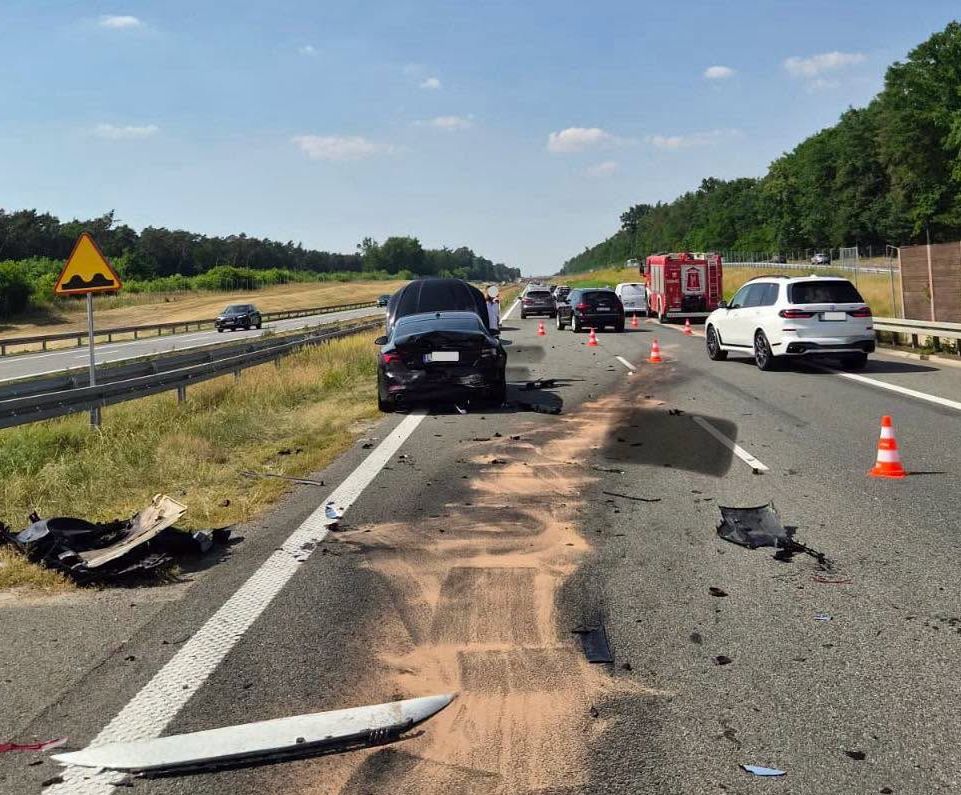 Na drodze widoczny wypadek samochodowy z uszkodzonym czarnym samochodem oraz rozrzuconymi fragmentami pojazdów. W tle widać samochody przejeżdżające obok, a także pojazd straży pożarnej.