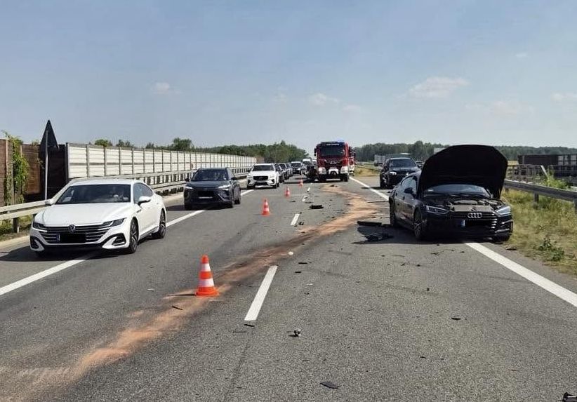 Czarne auto z otwartą maską stoi na jezdni, otoczone przez samochody i pachołki drogowe, na tle nieba.