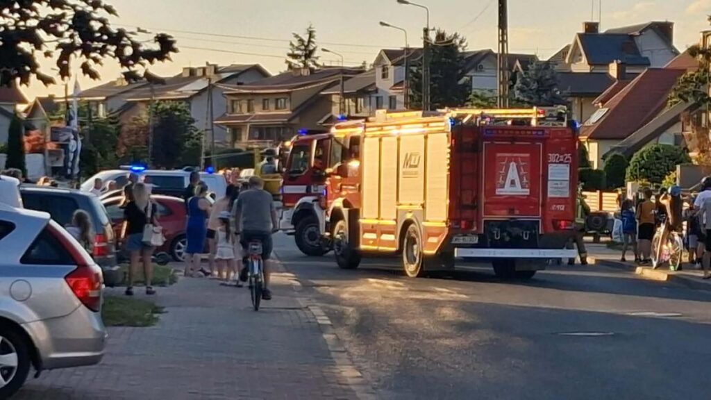 Grupa ludzi obserwuje działania służb ratunkowych przy strażackim wozie na ulicy w osiedlowej okolicy.