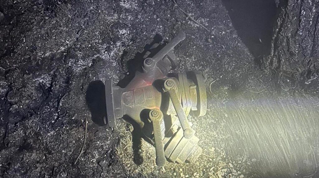 Uszkodzony element mechaniczny leży na ziemi w zniszczonym terenie, otoczony popiołem i resztkami roślinności.