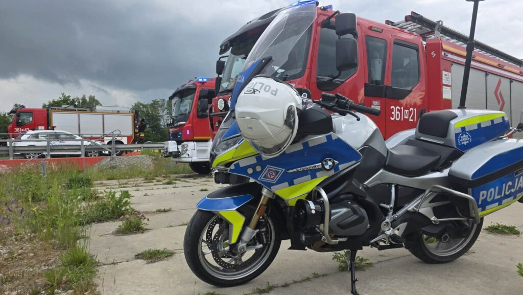 Motocykl policyjny w niebiesko-żółtej kolorystyce stoi na tle wozów strażackich na parkingu. Ciemne chmury w tle.
