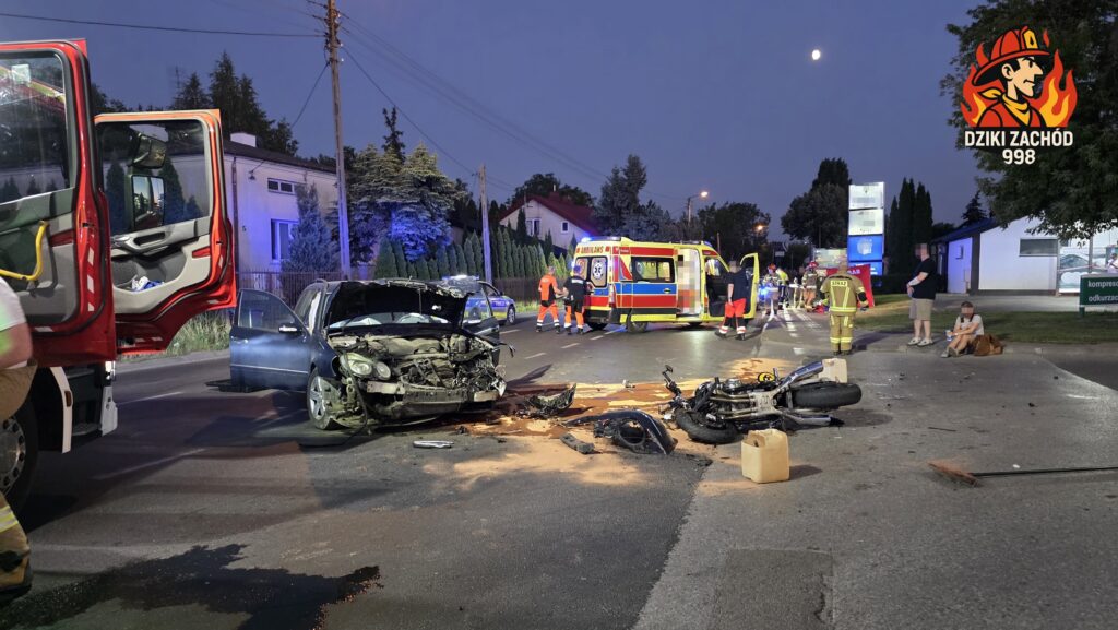 Na drodze leży rozbita ciężarówka oraz motocykl, a obok pracują ratownicy medyczni i strażacy. W tle widoczne są pojazdy służb ratunkowych.