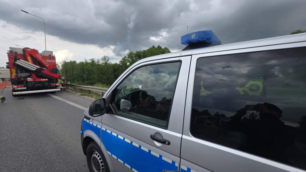 Radiowóz policyjny z niebieską sygnalizacją zatrzymany obok drogi, w tle widać czerwoną ciężarówkę z wysięgnikiem.