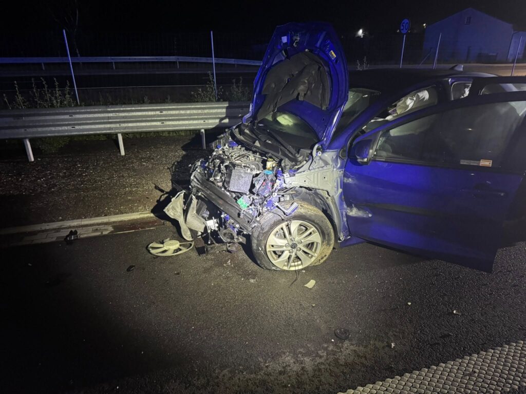 Zniszczony niebieski samochód osobowy z otwartą maską stoi na drodze, obok metalowej bariery ochronnej.
