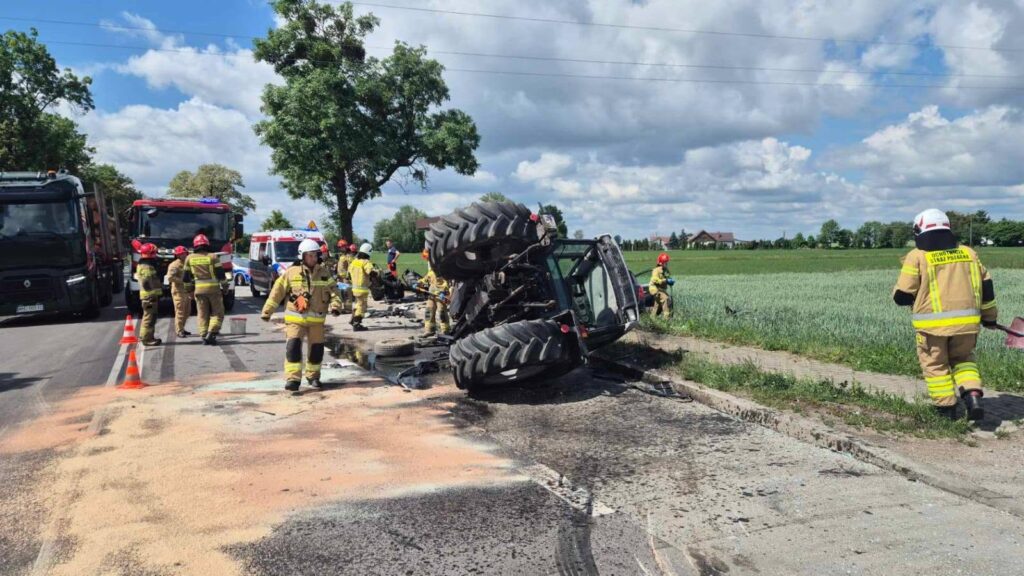 Strażacy w kaskach i mundurach usuwają skutki przewróconego ciągnika na drodze, obok pojazdów ratunkowych.
