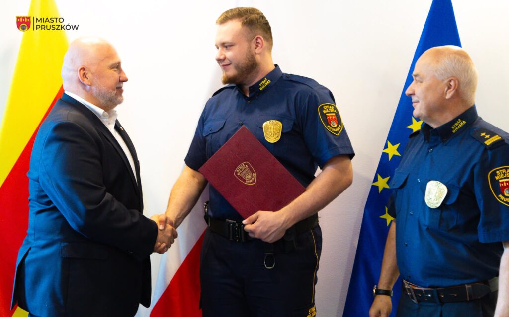 Mężczyzna w garniturze uścisnął dłoń strażnikowi miejskiego w mundurze, trzymającemu akt w czerwonej okładce.