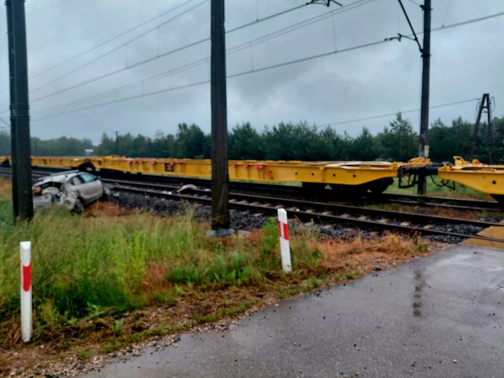 Samochód osobowy stoi na poboczu drogi obok torów kolejowych, w tle widoczne są żółte wagony towarowe.