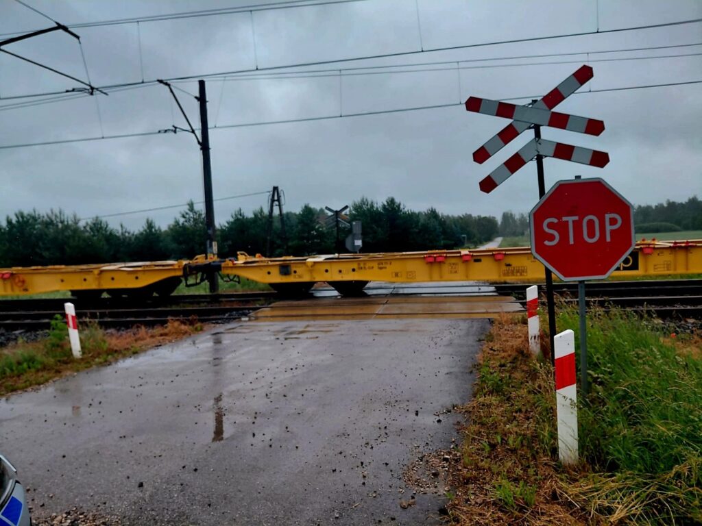 Na przejeździe kolejowym widoczne są żółte podkłady, znak stop oraz sygnalizacja ostrzegawcza. W tle zieleń i pochmurne niebo.