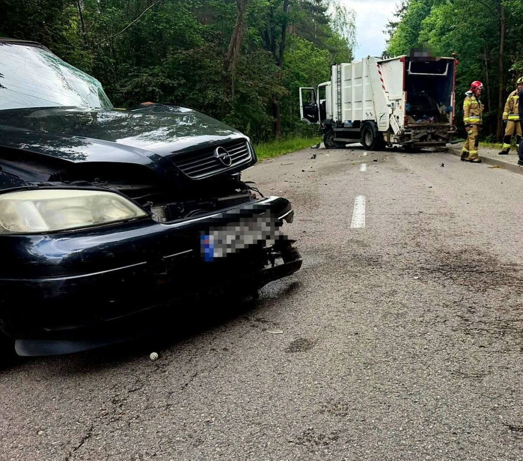 Zniszczony samochód osobowy stoi na drodze obok uszkodzonego pojazdu ciężarowego, w tle pracują strażacy.