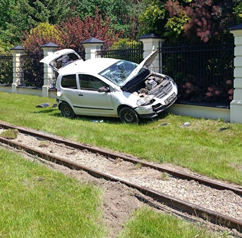 Biały samochód osobowy z otwartą maską stoi na trawie obok torów kolejowych, w otoczeniu gęstej roślinności.