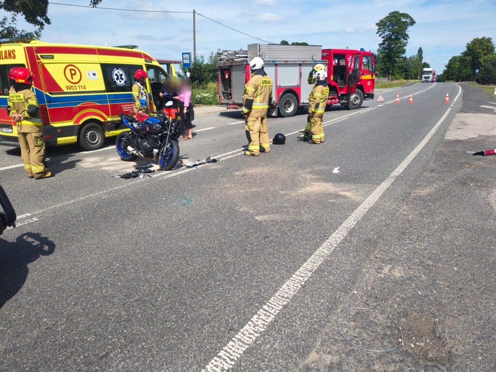Grupa strażaków w mundurach interweniuje na drodze, obok uszkodzonego motocykla i karetki pogotowia.