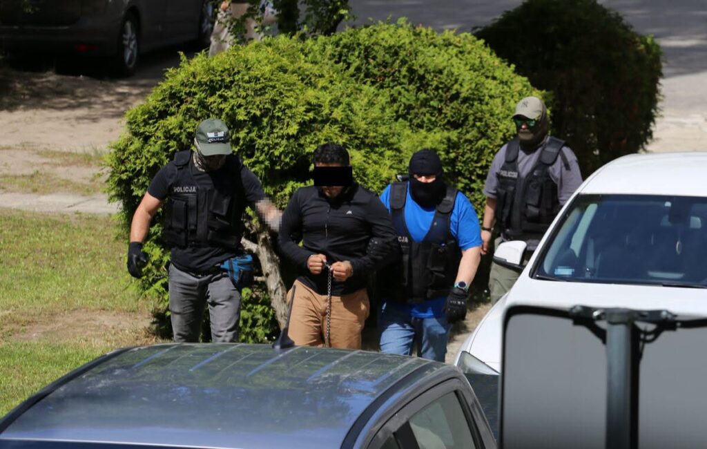 Czterej funkcjonariusze policji prowadzą podejrzanego, który ma założone kajdanki, w otoczeniu zielonych krzewów i zaparkowanych samochodów.