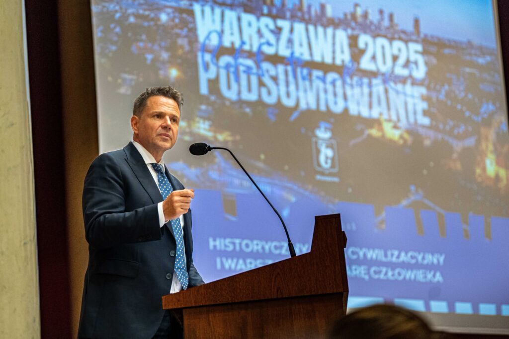 Mężczyzna w garniturze przemawia przy mównicy, w tle projektor z napisem "Warszawa 2025 - Podsumowanie".