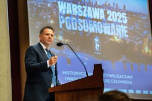 Mężczyzna w garniturze przemawia przy mównicy, w tle projektor z napisem "Warszawa 2025 - Podsumowanie".