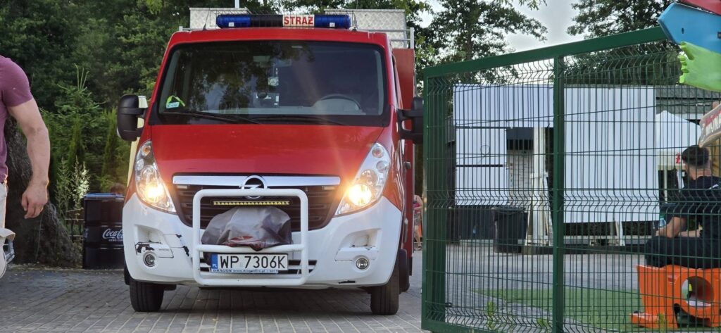 Czerwony wóz strażacki z włączonymi sygnałami świetlnymi podjeżdża do ogrodzonego terenu.