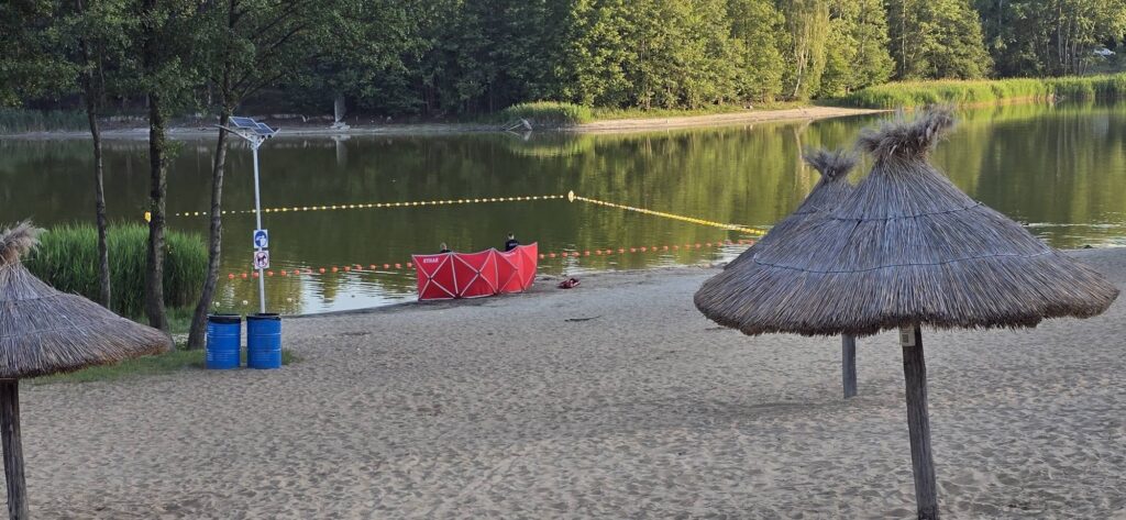 Dwie osoby siedzą w czerwonej łodzi na brzegu stawu, otoczonej zielenią i plażą z piaskiem.