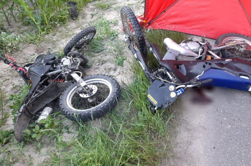 Dwa przewrócone motocykle terenowe leżą na trawie obok drogi, w otoczeniu zieleni i piasku.