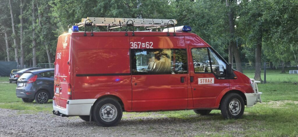 Czerwony samochód strażacki zaparkowany na trawiastej przestrzeni otoczonej drzewami.