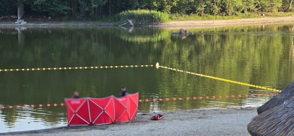 Dwóch mężczyzn siedzących za czerwonym parawanem na plaży przy jeziorze, z widoczną liną zabezpieczającą.