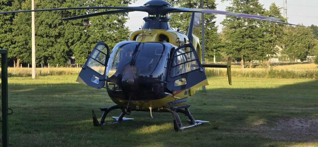 Helikopter z otwartymi drzwiami ląduje na trawiastej przestrzeni otoczonej drzewami.