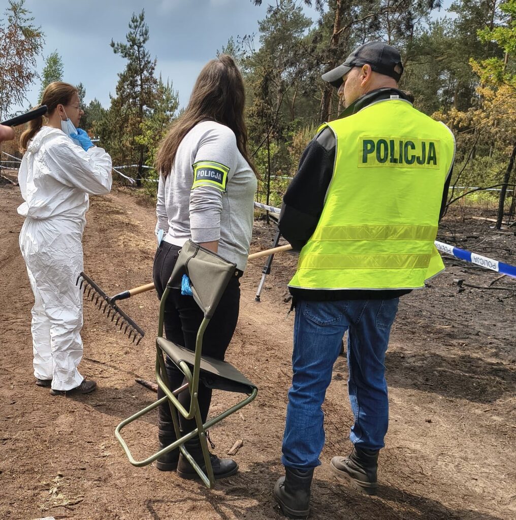 Kobieta w białym kombinezonie i mężczyzna w kamizelce z napisem "POLICJA" rozmawiają na terenie pożaru lasu.