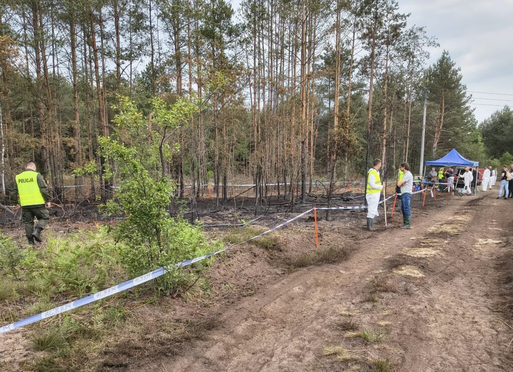 Policjant w kamizelce odblaskowej przeszukuje teren po pożarze, w tle zespół ludzi w białych kombinezonach.