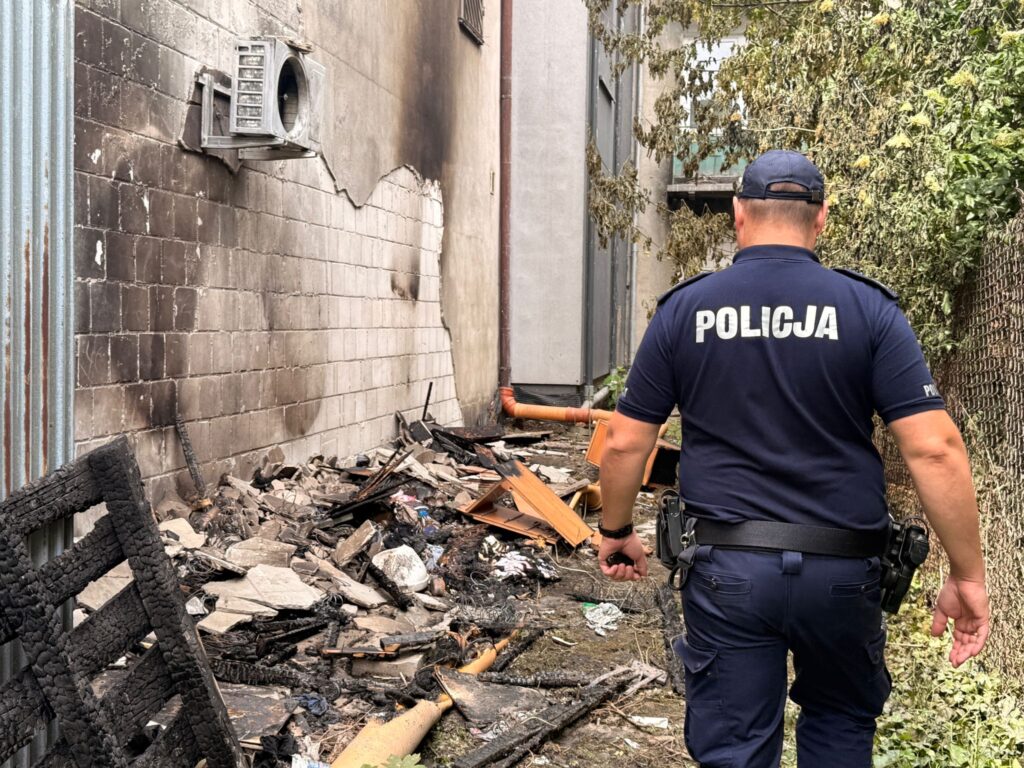 Policjant w mundurze przechadza się obok zniszczonego terenu z gruzem i popiołem, przy ścianie budynku.