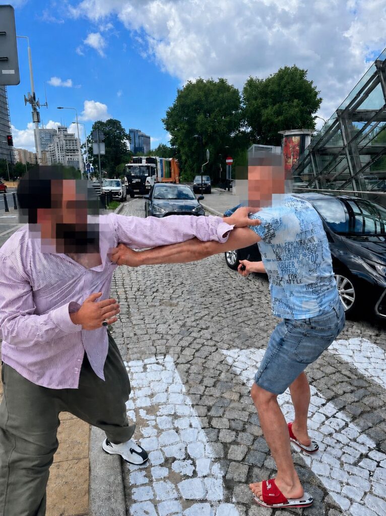 Dwaj mężczyźni szarpią się na ulicy w miejskim otoczeniu, w tle widoczne samochody i nowoczesne budynki.