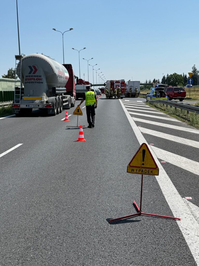 Policjant kierujący ruchem na drodze, obok ciężarówki i wozów strażackich, oznaczenia „WYPADEK” w tle.