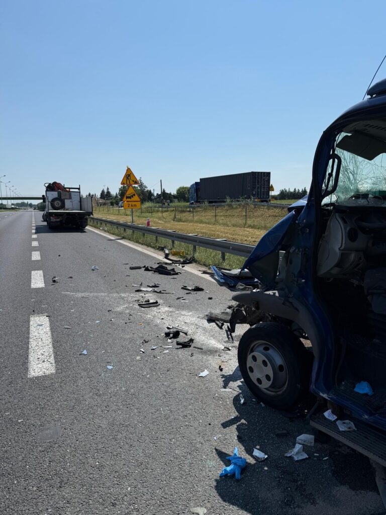 Rozbite auto na drodze, wokół leżą szczątki pojazdu oraz resztki papierów. W tle znak ostrzegawczy o robach drogowych.