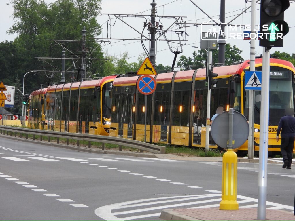 Tramwaj w kolorze żółto-czerwonym zatrzymany na przystanku przy ruchliwej ulicy, obok znaków drogowych.