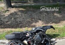 Motocykl leżący na poboczu drogi, z uszkodzoną konstrukcją i wyciekającymi płynami. W tle widoczna zieleń.