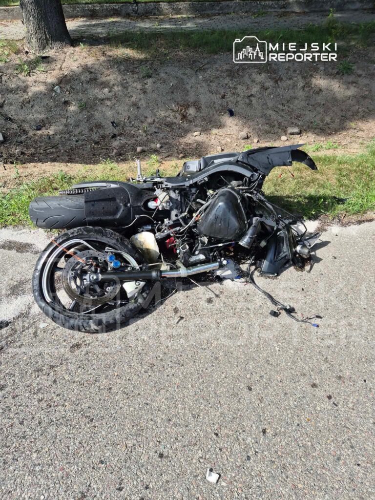 Motocykl leżący na poboczu drogi, z uszkodzoną konstrukcją i wyciekającymi płynami. W tle widoczna zieleń.
